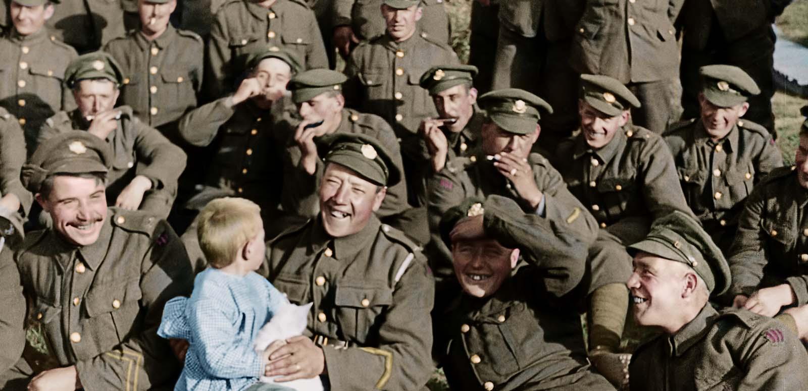 Three rows of British soldiers in uniform relaxing on the grass. One at the front is smiling and holding a small child.