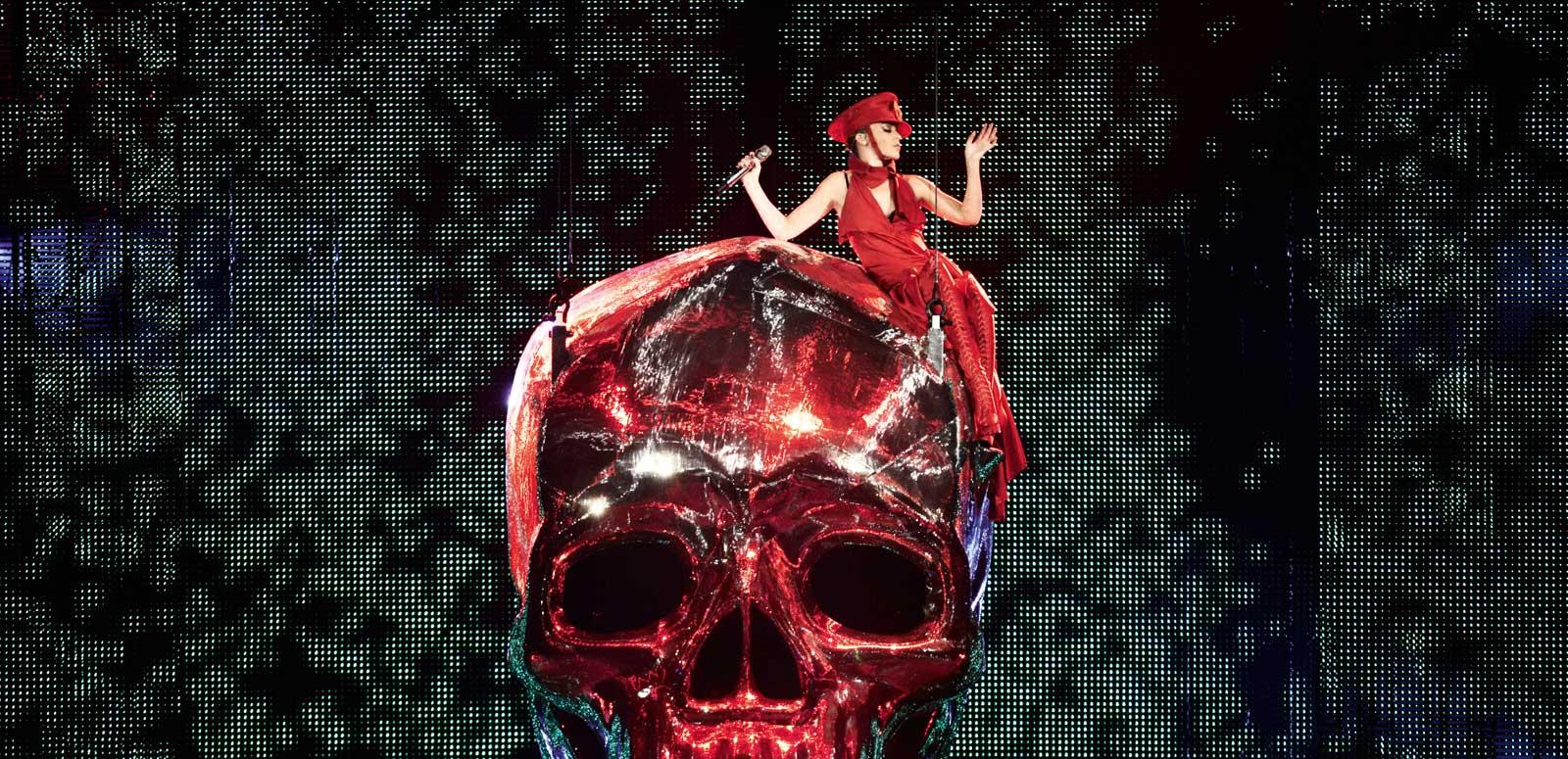 Kylie Minogue, dressed entirely in red, is sitting on top of a giant red shiny skull prop at one of her live shows. 