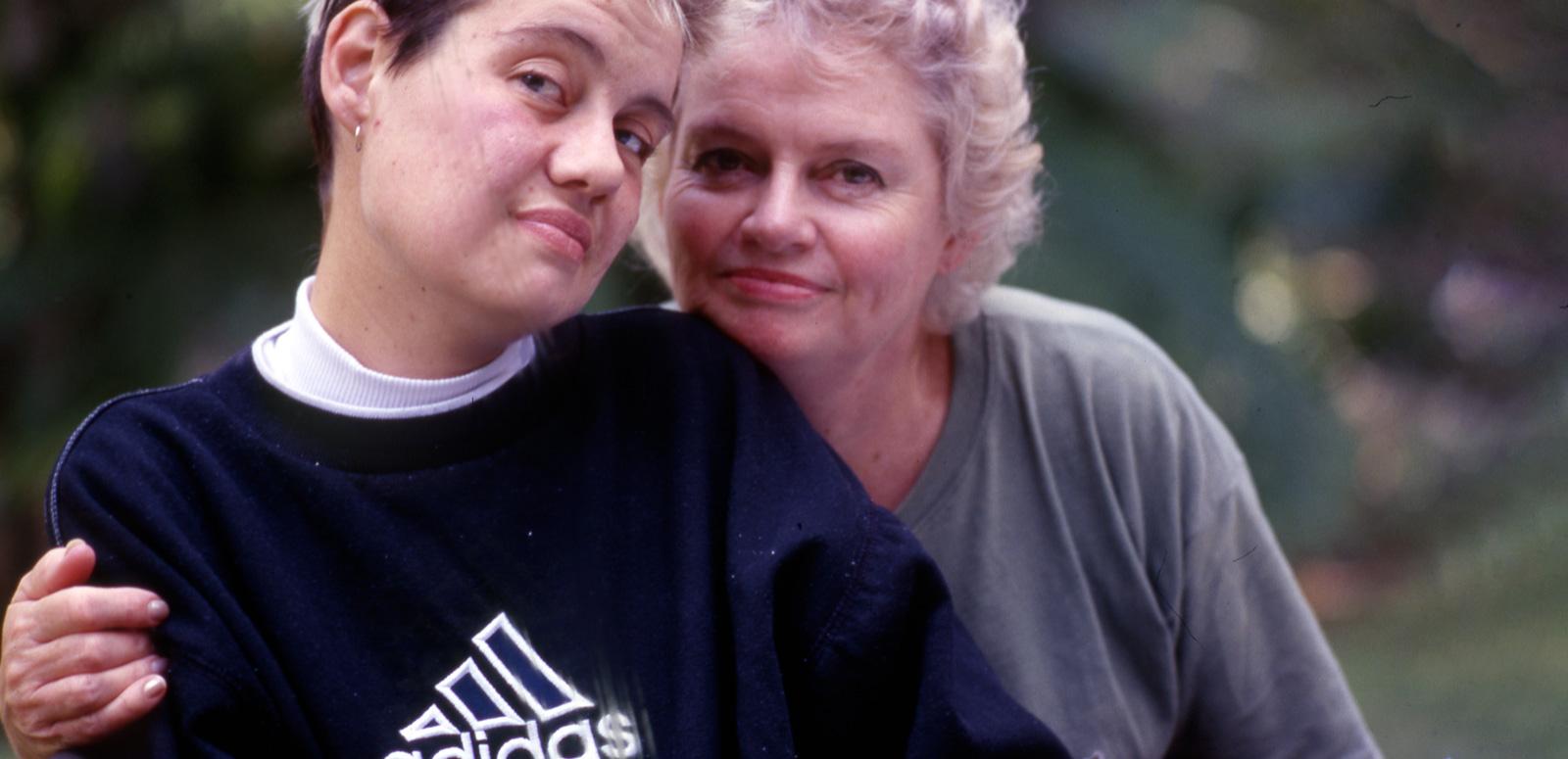 A young woman with short blond hair, wearing a black jumper is embraced by an older blond lady. They are looking at the camera.