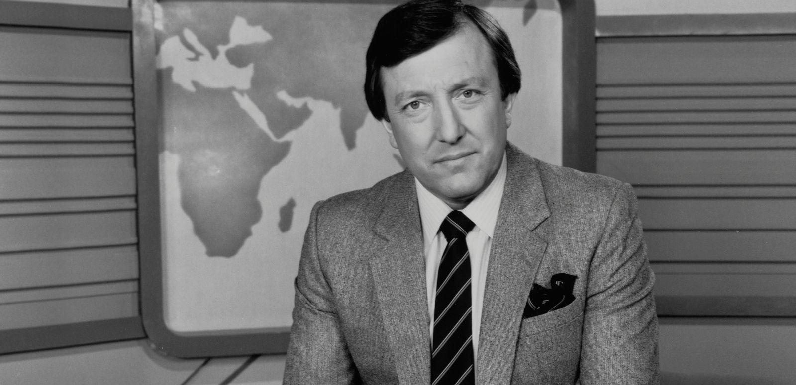 Journalist Jim Waley is wearing a suit and tie in this black and white photo. He is sitting in a studio with a backdrop showing a map of the world.