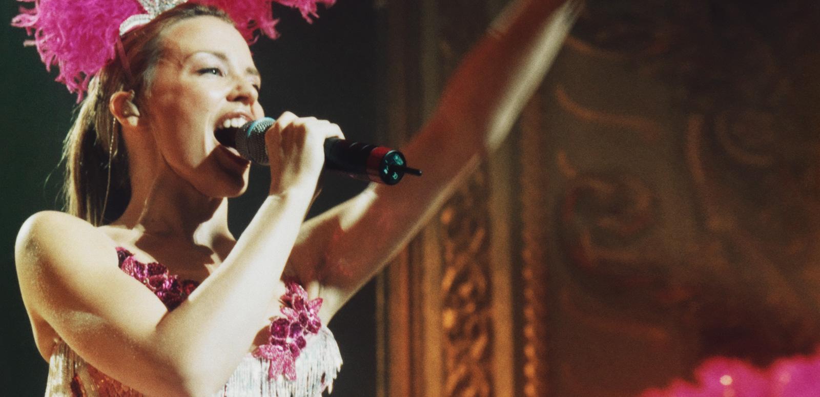 Kylie Minogue on stage in a pink showgirl costume singing into a microphone.