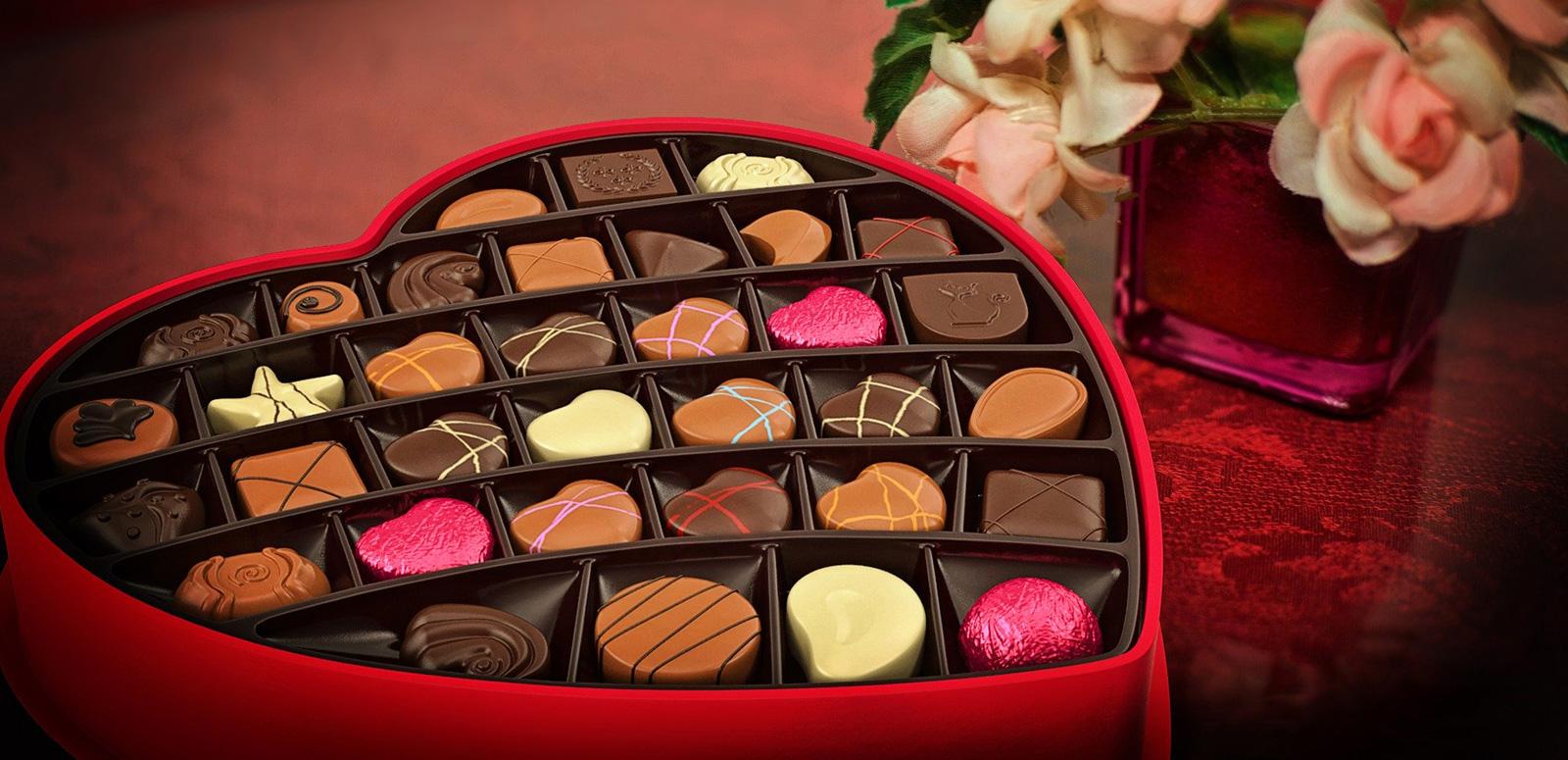 A heart shaped box of assorted chocolates with a small vase of pink roses in the background