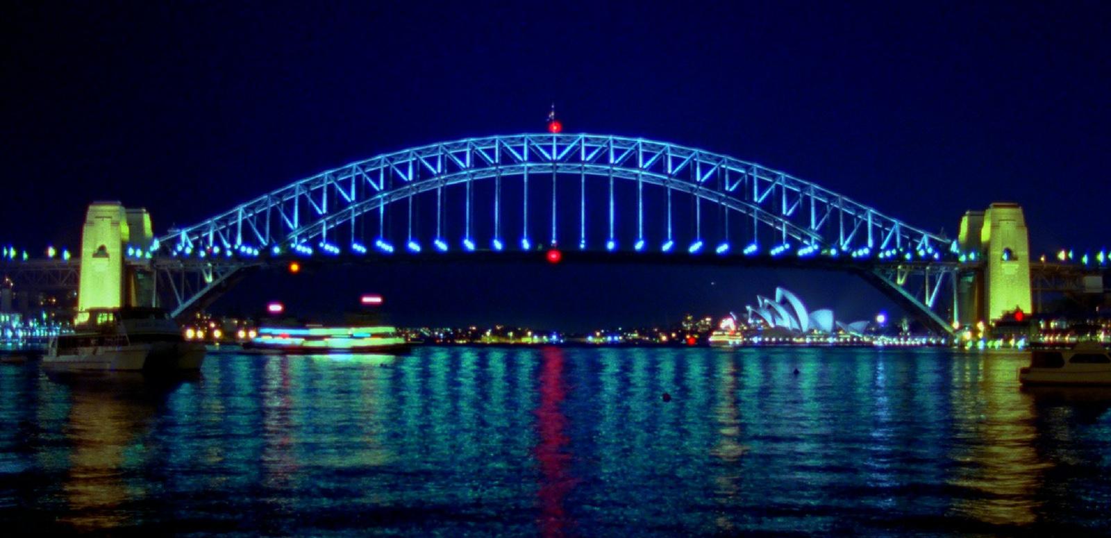 A nightime photo of the Sydney Harbour Bridge showing it all lit up. The bridge arch is blue with red lights at the top and bottom centre. The pylons are lit yellow. The water shows the reflections of the passing ferry and the Sydney Opera House.
