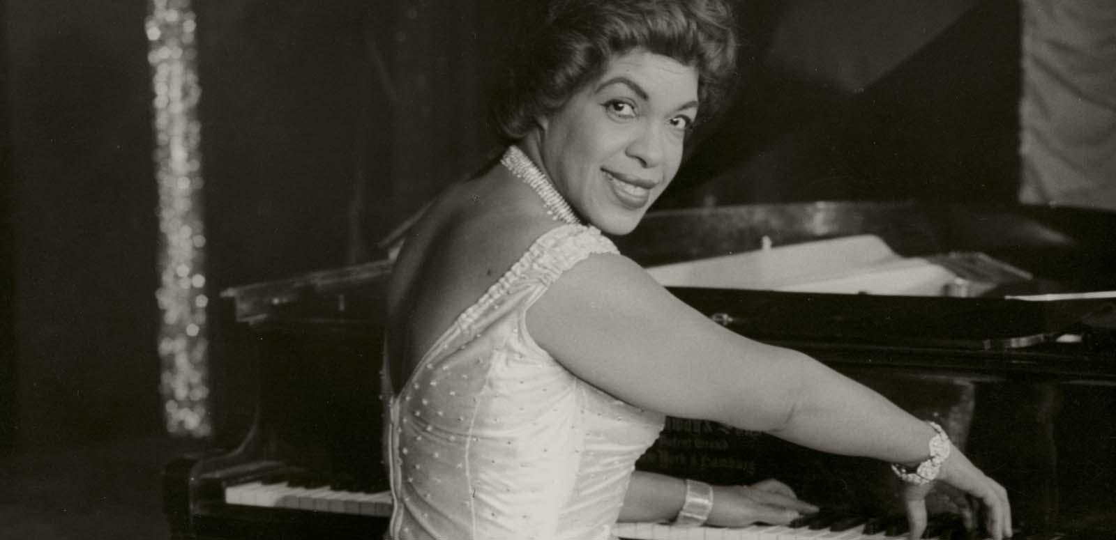Winifred Atwell seated at the piano turns to smile at the camera