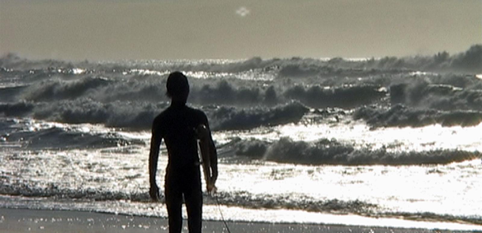 Silhouette of a man with his back to camera walking into the surf carrying a surfboard under one arm.