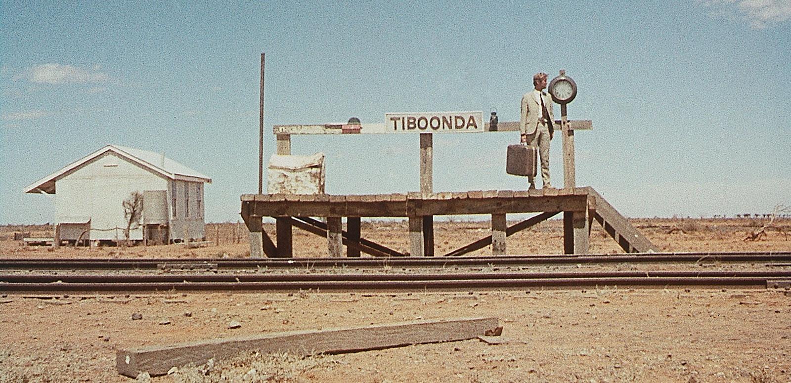 A man carrying a suitcase and wearing a light brown suit waits on a small outback railway platform