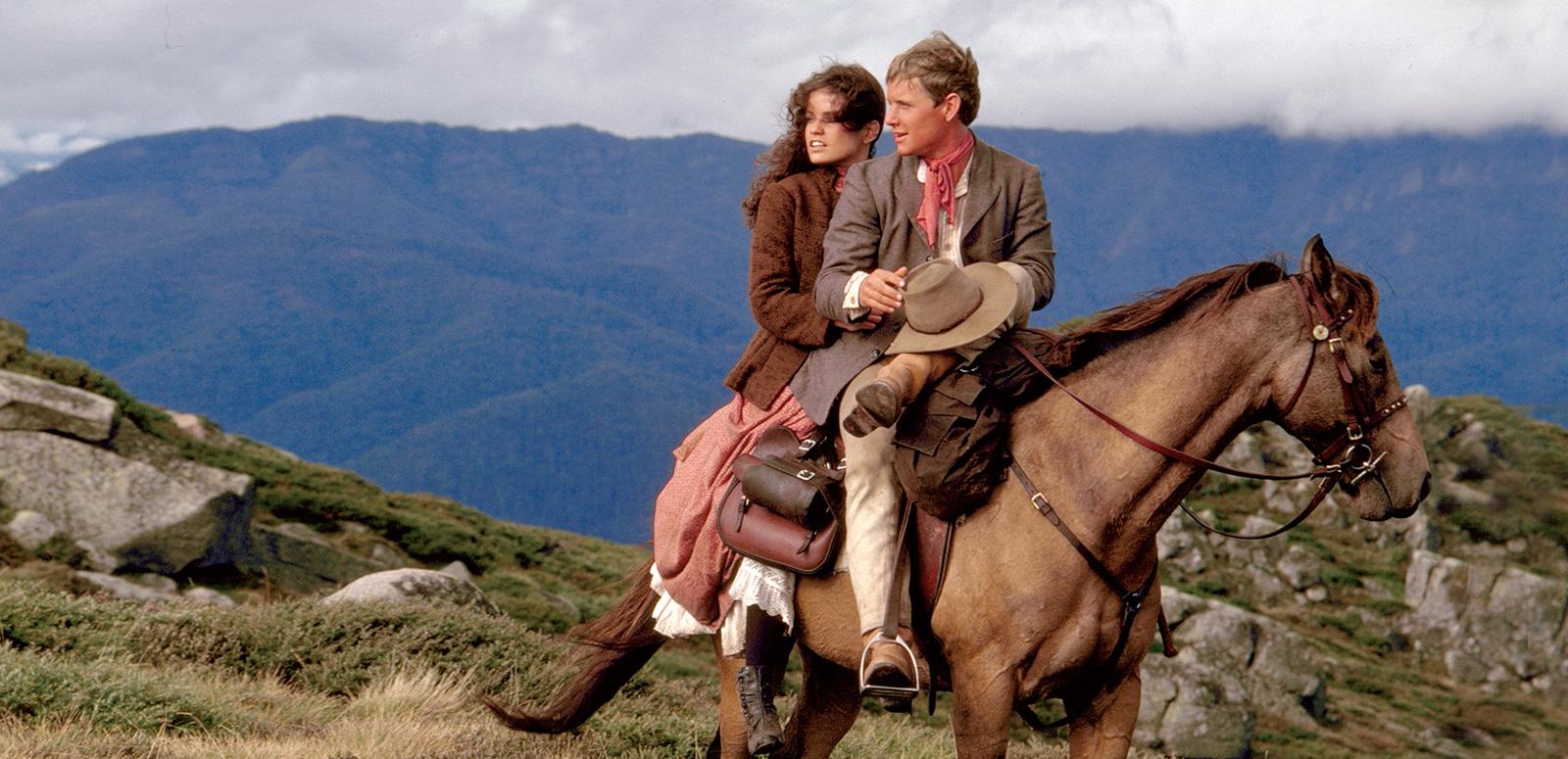 Sigrid Thornton and Tom Burlinson on a chestnut coloured horse with mountains in the background in a scene from 'The Man From Snowy River'.