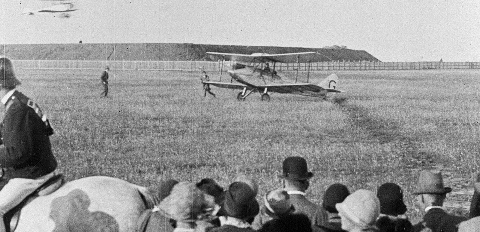 An early 20th century biplane lands in a field, watched by a crowd of spectators