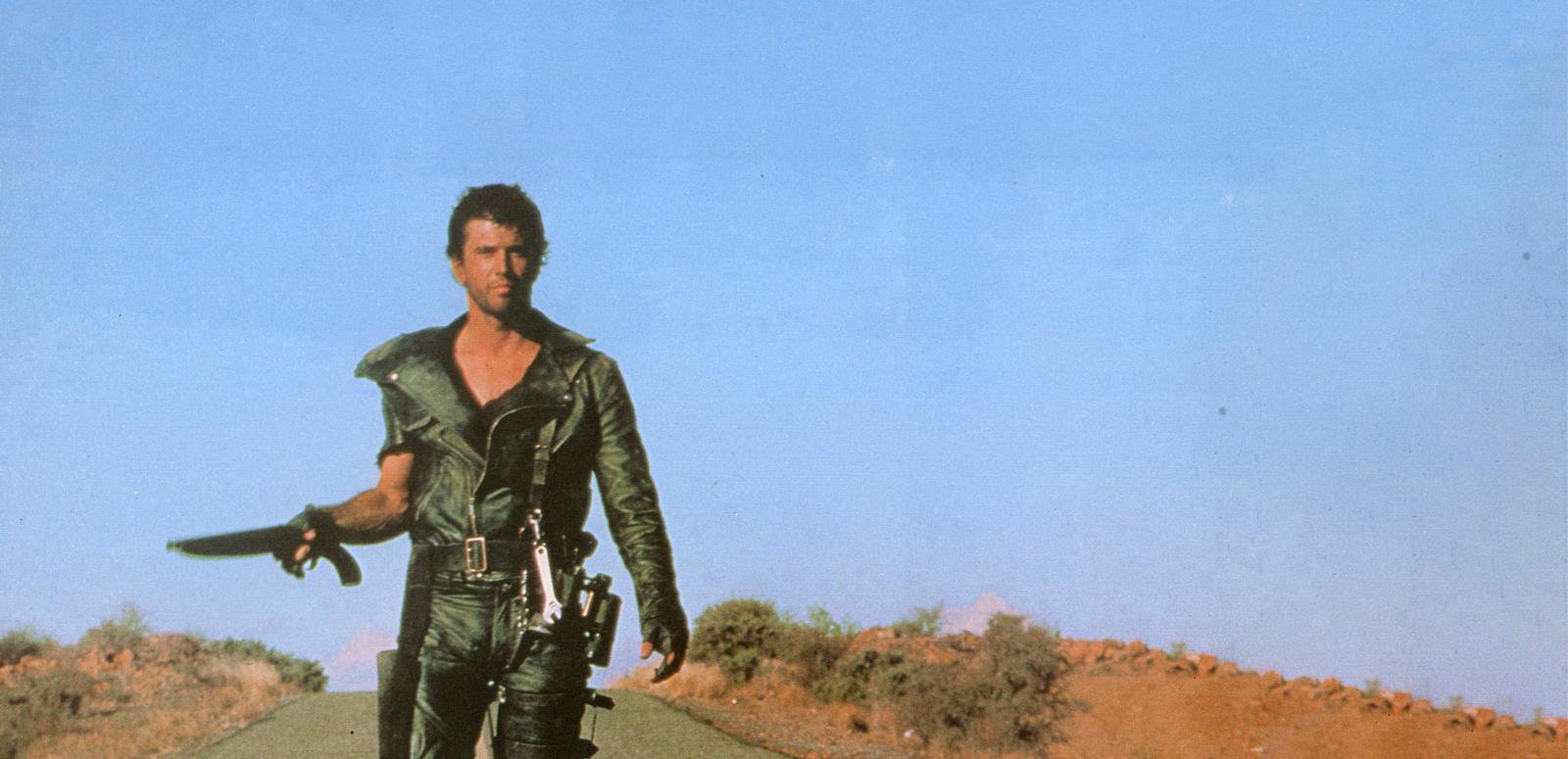 Mad Max walking resolutely down the middle of a desert highway, gun in hand