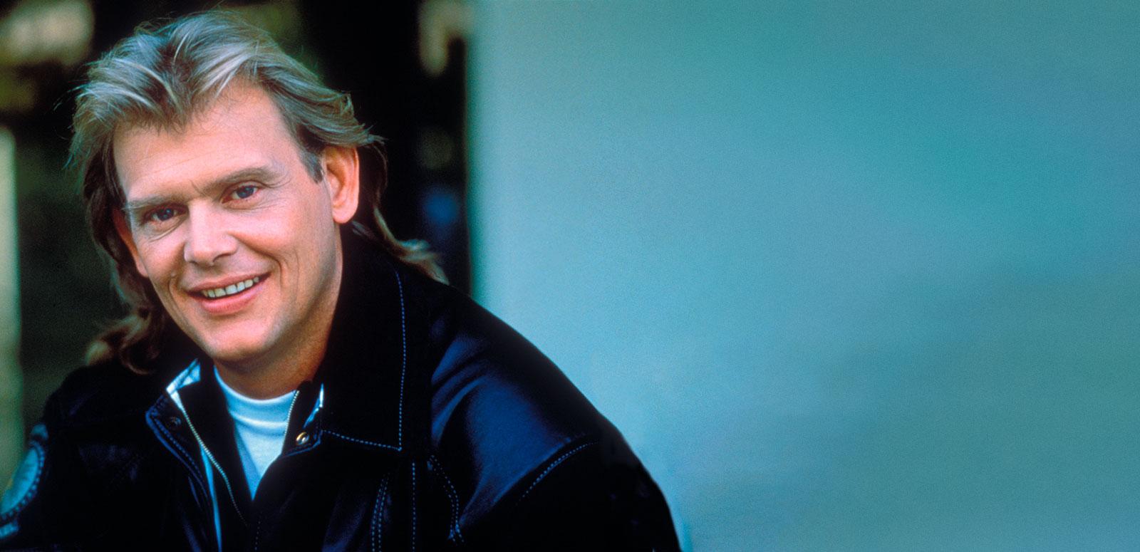 Singer John Farnham, circa late 1980s wearing a black jacket, pictured from the shoulders up. He has his head tilted slightly towards the camera and is smiling.