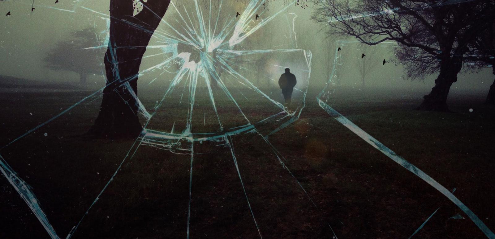 View from a window that has been shattered showing a dark, shadowy figure of a man walking through a grassy field between some trees.
