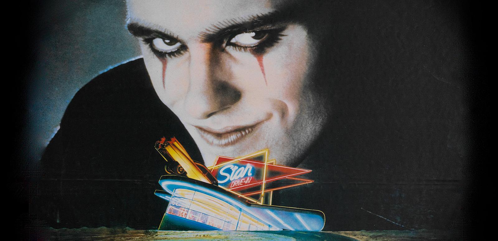 A close-up of a menacing looking man with dark eye make-up and an evil grin. His head looms over a smaller image of a neon-lit drive-in cinema and empty road.