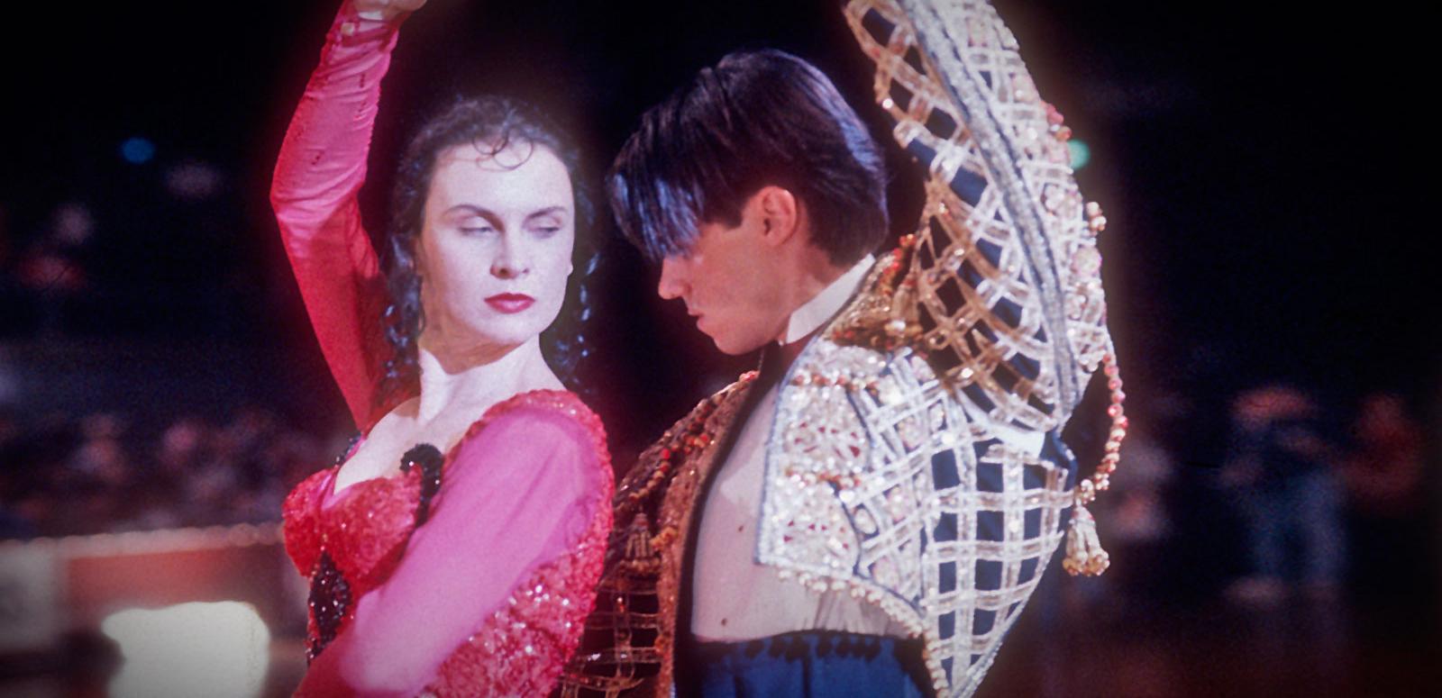 Tara Morice and Paul Mercurio in a dance pose in a scene from the film Strictly Ballroom. She is wearing a red and black beaded and ruffled flamenco style dress and he is wearing a spanish style gold and black bolero jacket. 