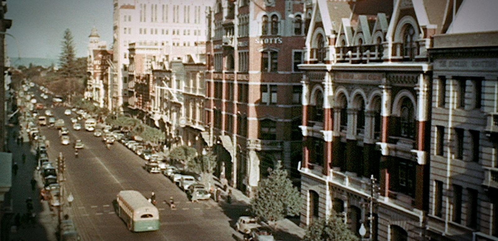 A street scene from Perth circa 1954. The street is lined with 3 and 4 story buildings and there are many cars parked on the side of the road, there are trees along the footpaths and a few pedestrians can be seen crossing the street.