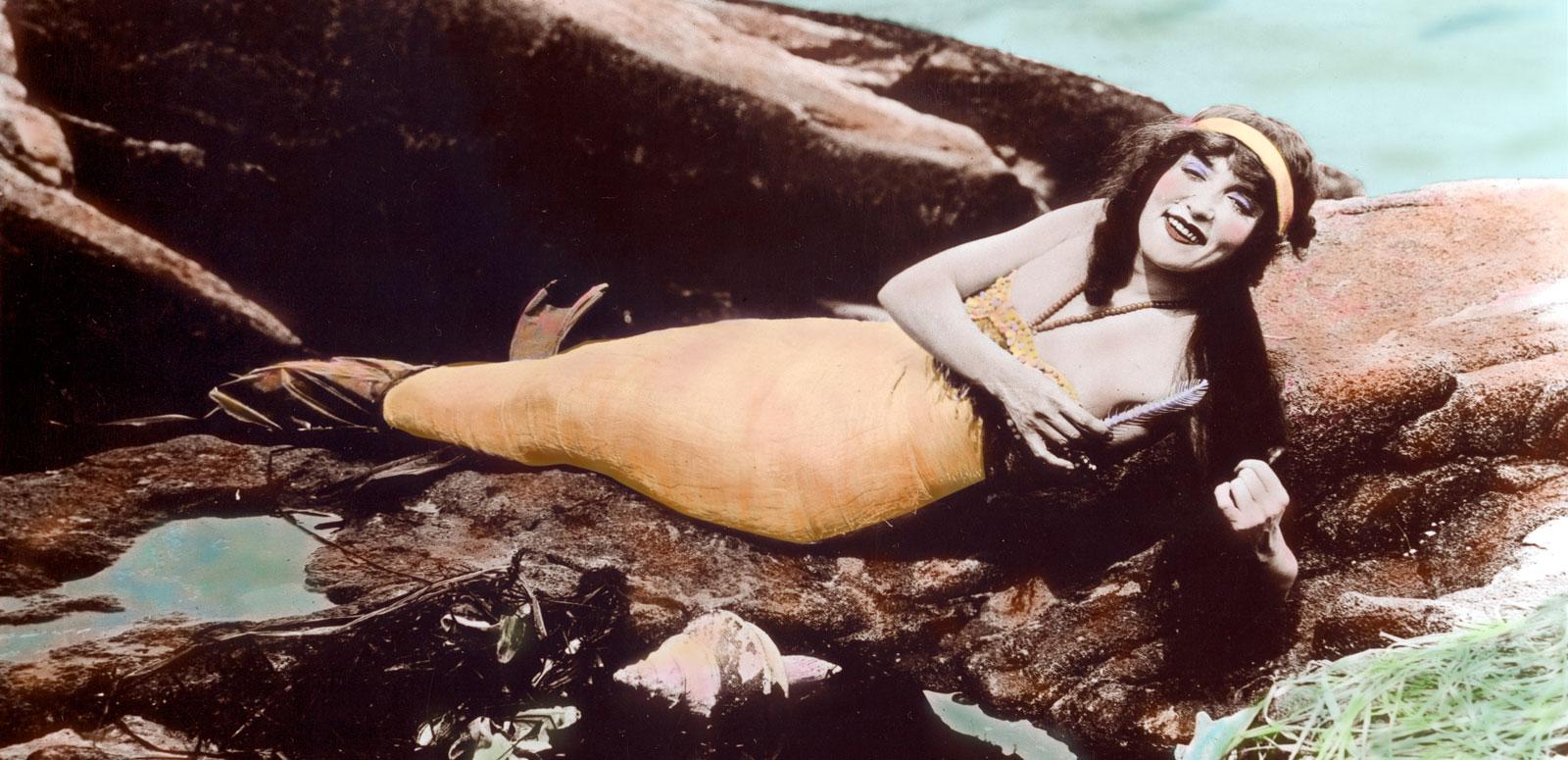 A hand-tinted photograph of Annette Kellerman wearing a mermaid costume and lying on rocks near the water.