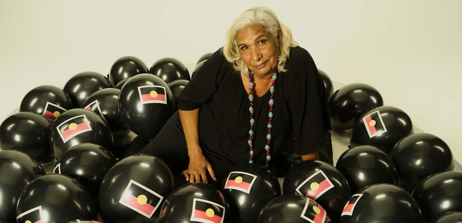 Trisha Morton-Thomas wearing a black dress and surrounded by black balloons with the Aboriginal flag on them
