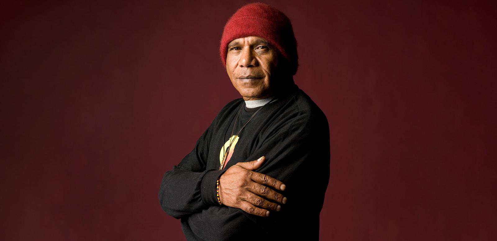 Archie Roach looks at the camera with his arms folded.