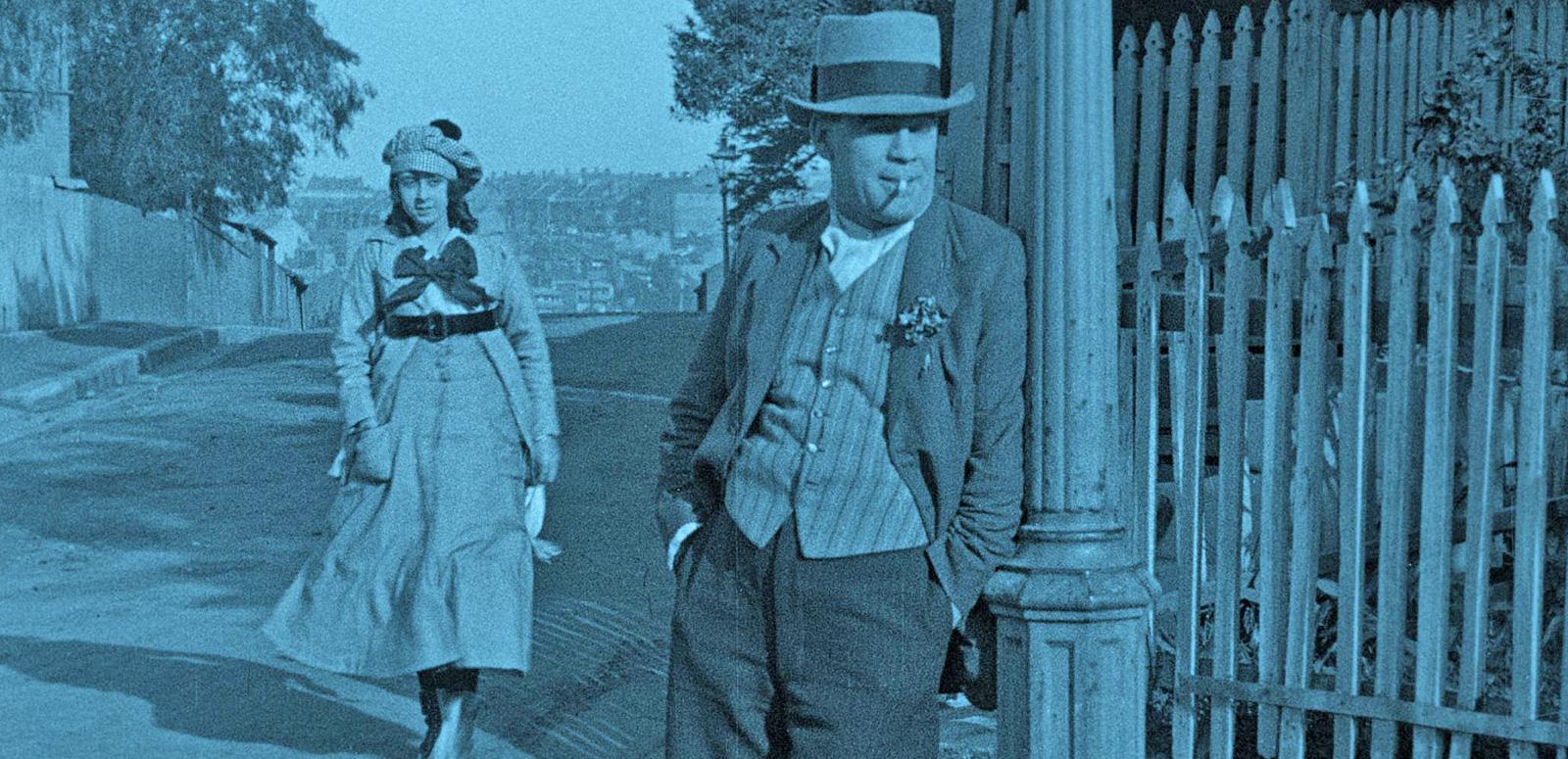 A scene from the film The Sentimental Block featuring Arthur Tauchert as 'The Bloke' leaning against a lamp post on a street corner Actress Lottie Lyell (Doreen) is walking up the street behind him.