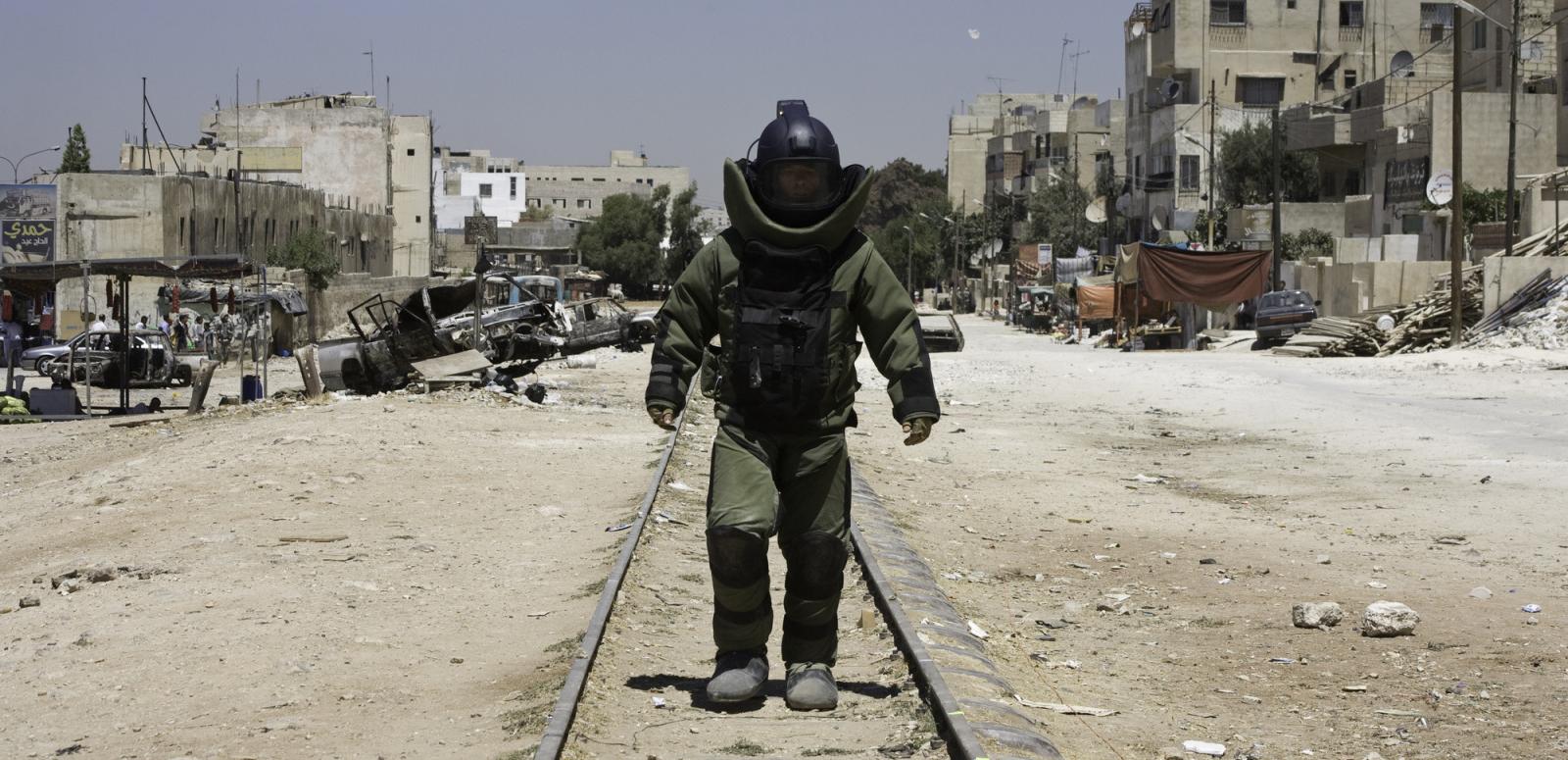 A lonely figure wearing bomb disposal protection walks toward the camera.