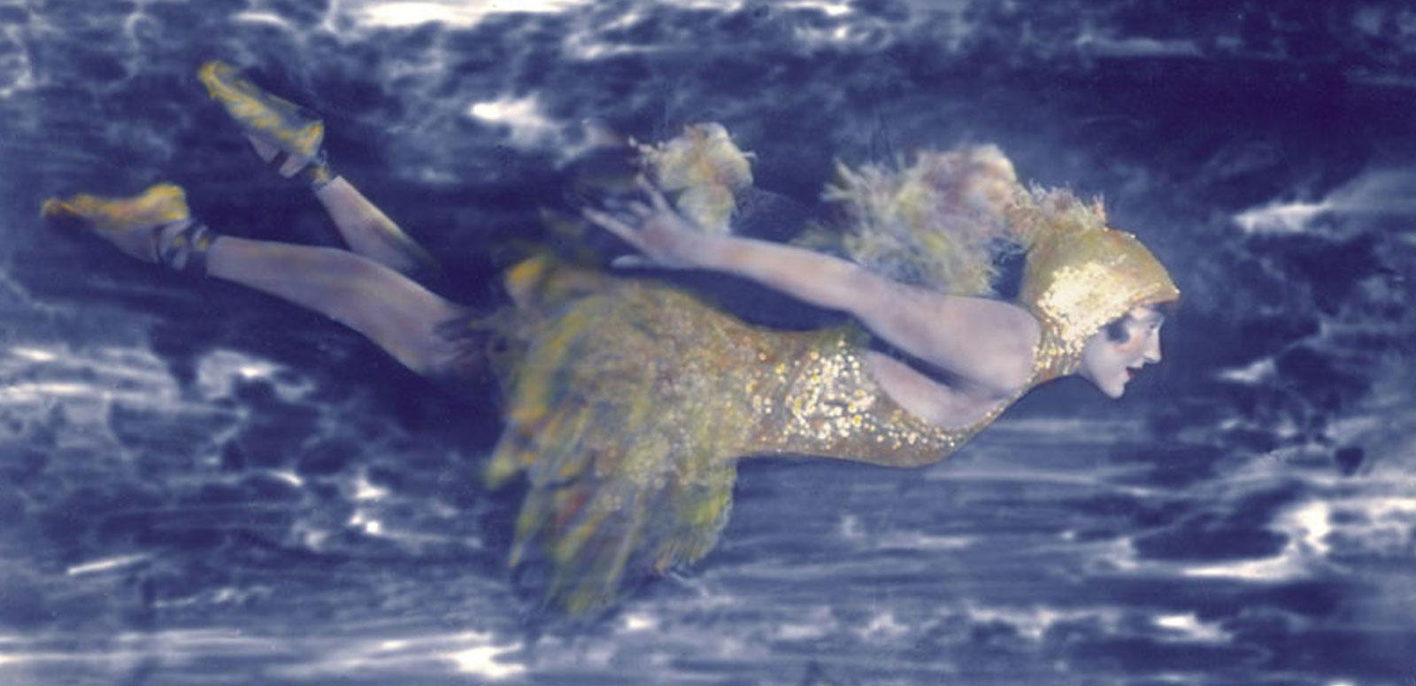 Hand-tinted photograph of Annette Kellerman swimming underwater wearing an elaborate costume including ballet slippers and head-dress.