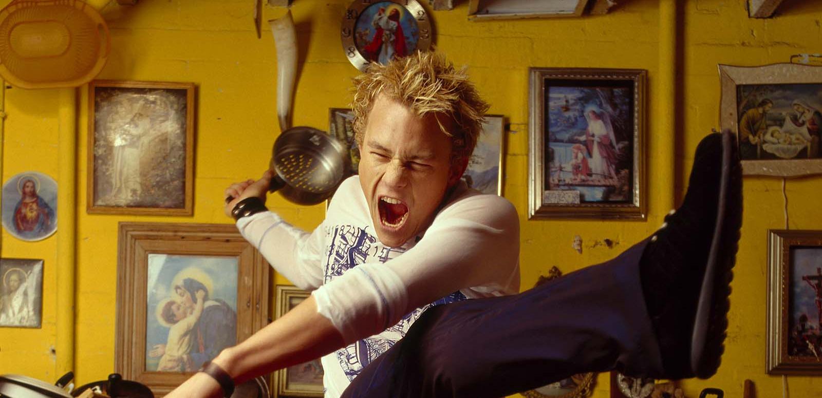 Actor Heath Ledger striking a dramatic pose with his arms out, one leg up in the air and his mouth open wide and eyes shut. He is in a yellow kitchen.