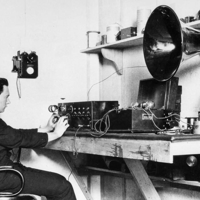 A man sitting on a desk with headphones on. There is radio and other audio equipment on the desk. c1920s