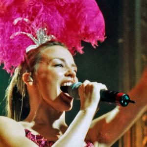 Kylie Minogue on stage in a pink showgirl costume singing into a microphone.