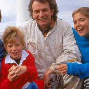 A family of four - two boys, a girl and their dad, kneeling with their arms around each other and smiling at the camera. You can see part of a lighthouse in the background. 