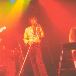 The 5 male members of 1970s rock band Sherbet performing on stage: a lead guitarist, bass guitarist, drummer and keyboards player with lead singer Daryl Braithwaite
