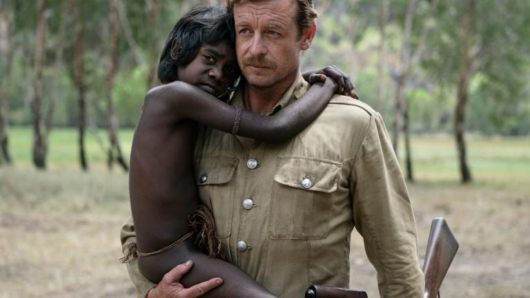 A white man in a uniform and holding a rifle is carrying a young Aboriginal boy in an outback setting.