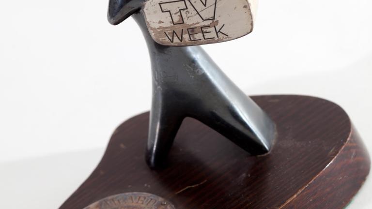 A TV Week Logie Award, made up of a small silver statuette in the shape of a man holding a television. The statuette sits on a round wooden base with a metal disc with words engraved on it. 