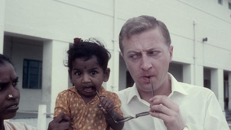 An Indian mother hold her child while the child and Graham Kennedy suck on the arms of Kennedy's glasses. Kennedy has a wry expression