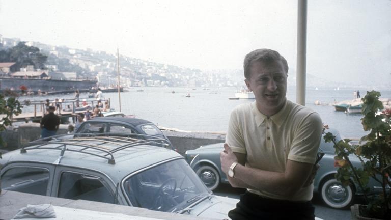 Graham Kennedy, arms folded, stands in front of cars at an Italian seaside town