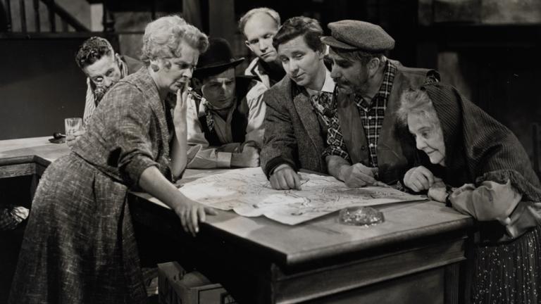 A group of people are crowded around a table in a still from a television production.