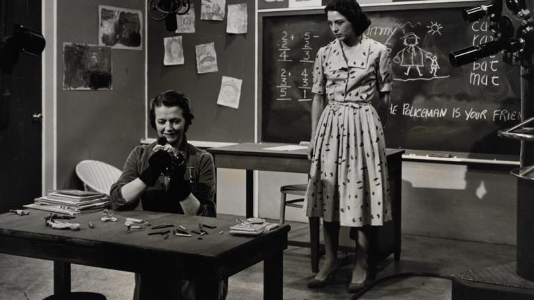 A production still of two women in a school room setting. A boom is visible in the background.