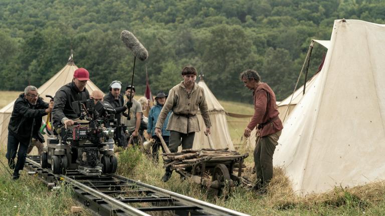 Behind the scenes shot of a cinematographer on a dolly filming a movie scene in a grassy field where a man in period costume is pushing a wheelbarrow.