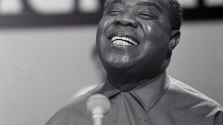 Close up, head and shoulders shot of jazz musician Louis Armstrong singing into a microphone with his eyes closed, circa 1964.
