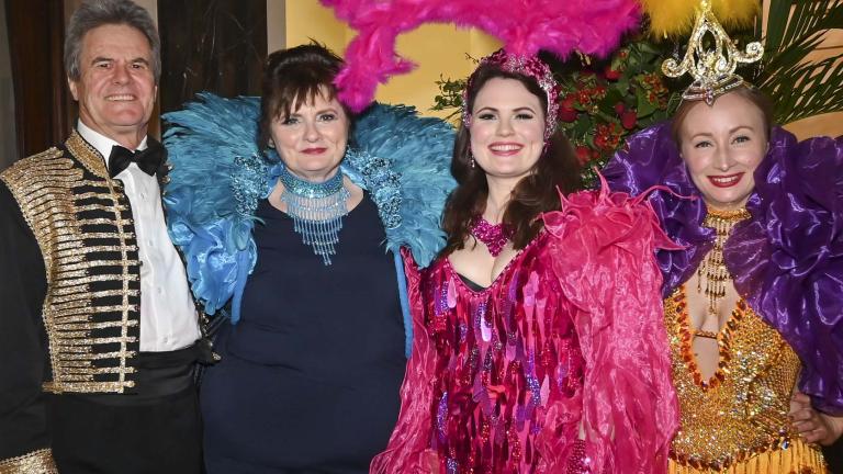 A man in a bolero jacket poses with three women in colouring sparkling outfits, including with elaborate feather headdresses