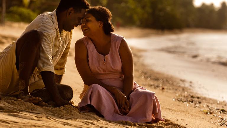 A First Nations man and woman sit together on a beach looking at each other and smiling in a scene from Mabo.