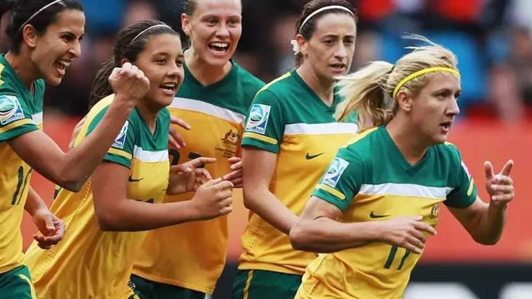 Five players from the Australian Women's Soccer team running side by side, they are smiling and look to be celebrating scoring a goal.