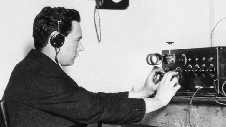 A white man wearing headphones twiddles with knobs on a 1920s crystal radio