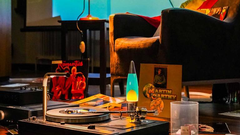 A table that has a turntable and records on it and a lava lamp. There is an armchair in the background. 