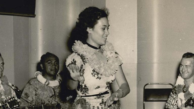 A Polynesian woman wearing a garland of flowers around her neck and dancing on stage in front of musicians during the recording of a 1950s radio talent show