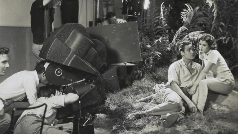 Charles Farrell and Mary Maguire on set sitting together in front of some ferns in front of a crew behind a large film camera