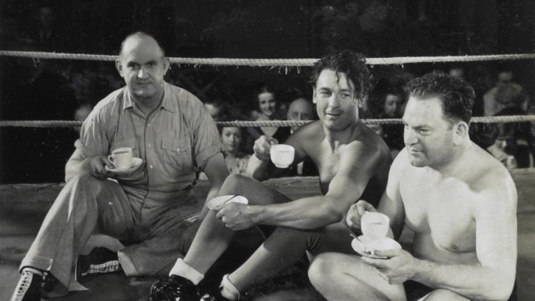 Referee Jim McMaster, Charles Farrell and professional wrestler Jack Clarke sitting in the middle of a wrestling ring drinking cups of tea
