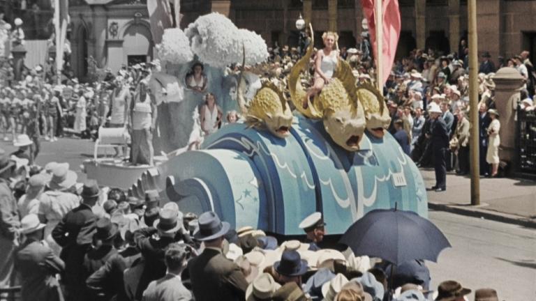 Colourised film still of floats in a parade.