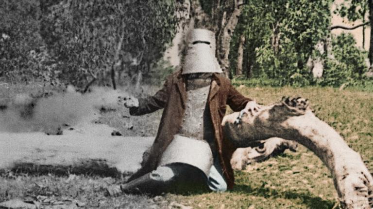Colourised film still of Ned Kelly in armour leaning against a dead tree trunk on the ground.