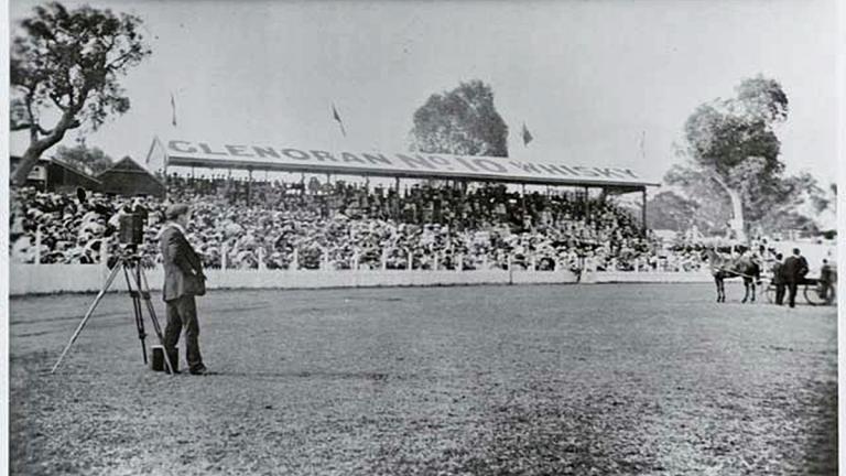 A man standing in an oval with a movie camera.  Behind him is a stand full of spectators and on the field is a group of men standing around a horse and carriage. c1900.