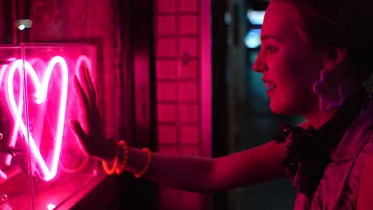 A woman places her hand against a shop window where a pink neon heart glows.