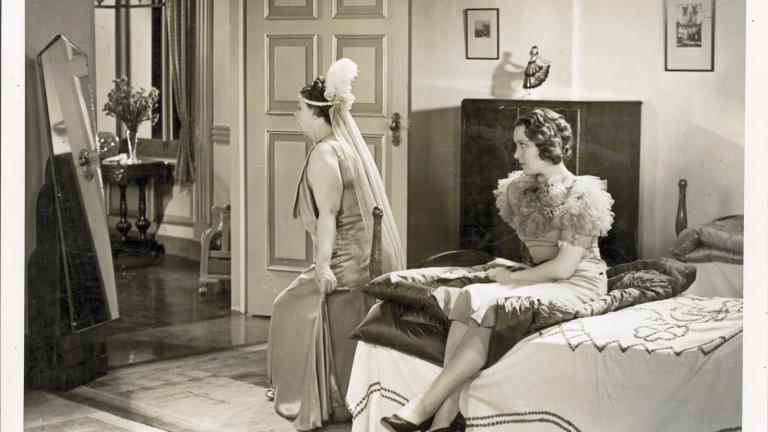 A woman wearing a ball gown and headdress looks in a long mirror while another young woman, seated on a bed, watches her.